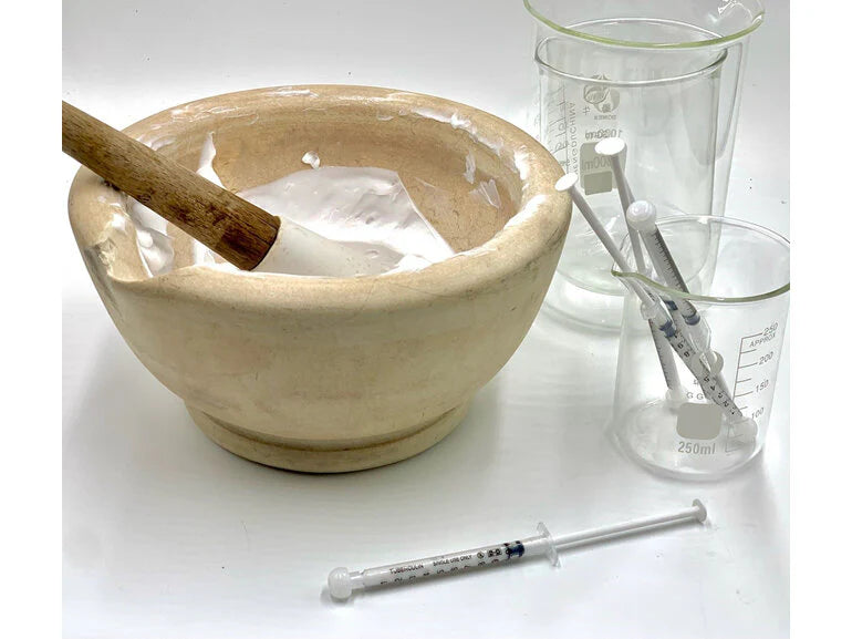 A beige mortar and pestle used for compounding with two glass beakers containing white spatulas and a syringe on a white surface, representing expert medication support for Bagga's Eyeliner Numbing Gel.