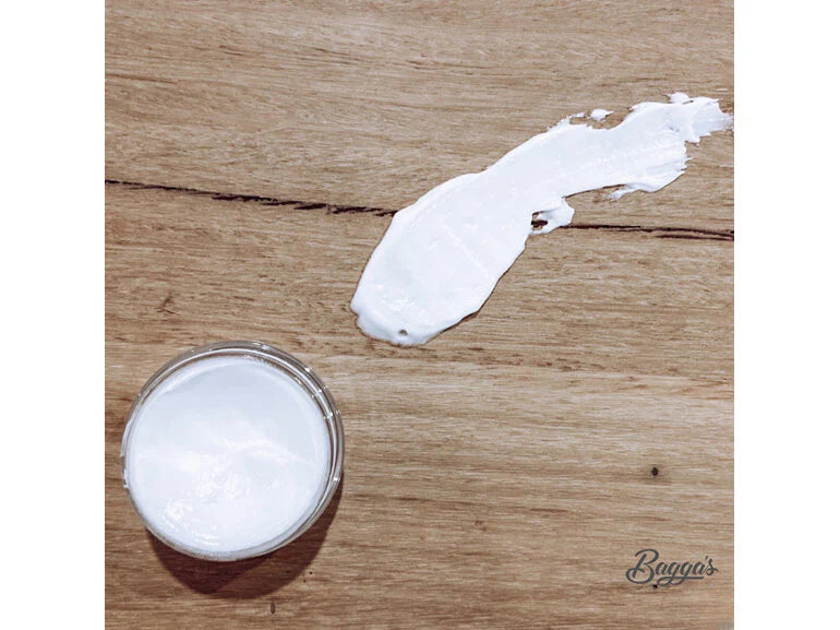 Open container and smear of white cream on wooden surface showing Bagga's Treatment Numbing Gel 10G texture.